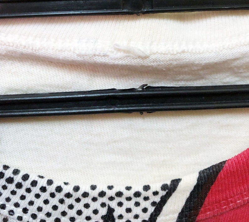 Puede incluir: Primer plano de una camiseta blanca con un estampado de lunares en blanco y negro en la parte inferior izquierda y una raya roja en la parte inferior derecha. La camiseta est&aacute; colgada de una percha negra.