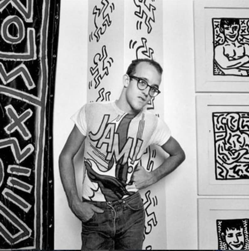 Puede incluir: Fotograf&iacute;a en blanco y negro de un hombre con gafas y una camiseta con la palabra "JAM!" en estilo c&oacute;mic. El fondo presenta obras de arte con figuras de palitos y dibujos enmarcados.