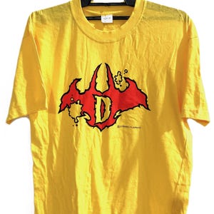 May include: Yellow t-shirt with a crew neck and short sleeves. The shirt features a red and black graphic design with the letter "D" in the center. The text "DYNAMIC PLANNING" is printed below the graphic.