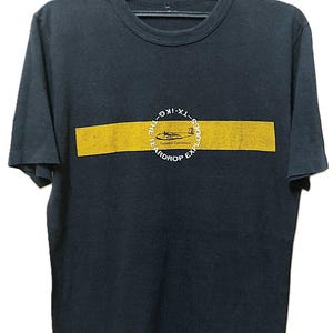 Può includere: T-shirt nero girocollo con una striscia orizzontale gialla sul petto. La striscia contiene una grafica bianca di un aereo e il testo "The Teardrop Explodes". La maglietta è appesa a una gruccia nera.