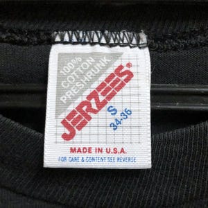 Op de afbeelding: Close-up van een zwarte t-shirtkraag met een wit label. Het label toont de merknaam "JERZEES" in rood, samen met details dat het shirt 100% katoen is, voorgekrompen en maat S heeft, met afmetingen van 86-91 cm. Het label vermeldt ook "MADE IN U.S.A."