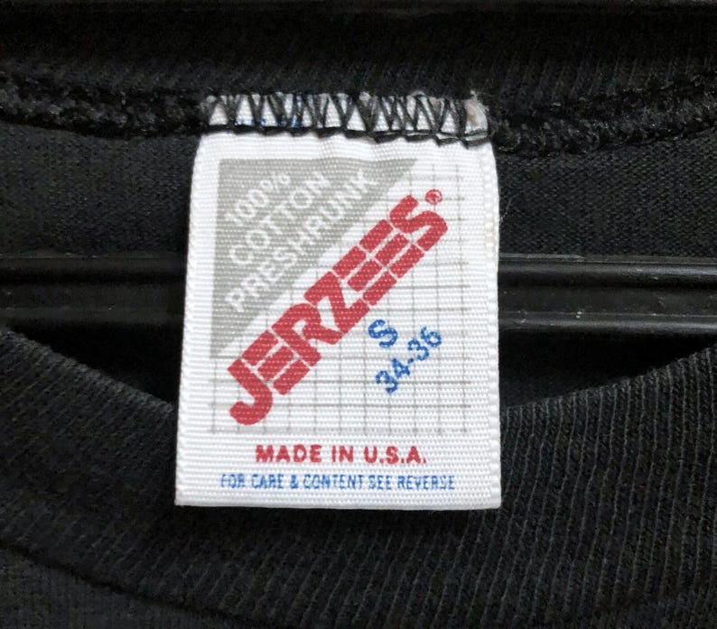 Op de afbeelding: Close-up van een zwarte t-shirtkraag met een wit label. Het label toont de merknaam "JERZEES" in rood, samen met details dat het shirt 100% katoen is, voorgekrompen en maat S heeft, met afmetingen van 86-91 cm. Het label vermeldt ook "MADE IN U.S.A."
