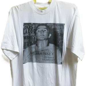 Può includere: T-shirt bianca con un'immagine in bianco e nero di una persona. Il nome "MORRISSEY" e "SOUTHPAW GRAMMAR" sono stampati sotto. La maglietta è appesa a una gruccia gialla.