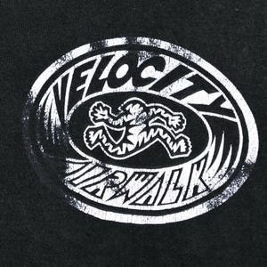 Op de afbeelding: Zwart t-shirt met een verweerde witte ovale afbeelding. De afbeelding toont de woorden "VELOCITY WALK" rond een gestileerde figuur in beweging. Het ontwerp heeft een vintage, versleten uitstraling.
