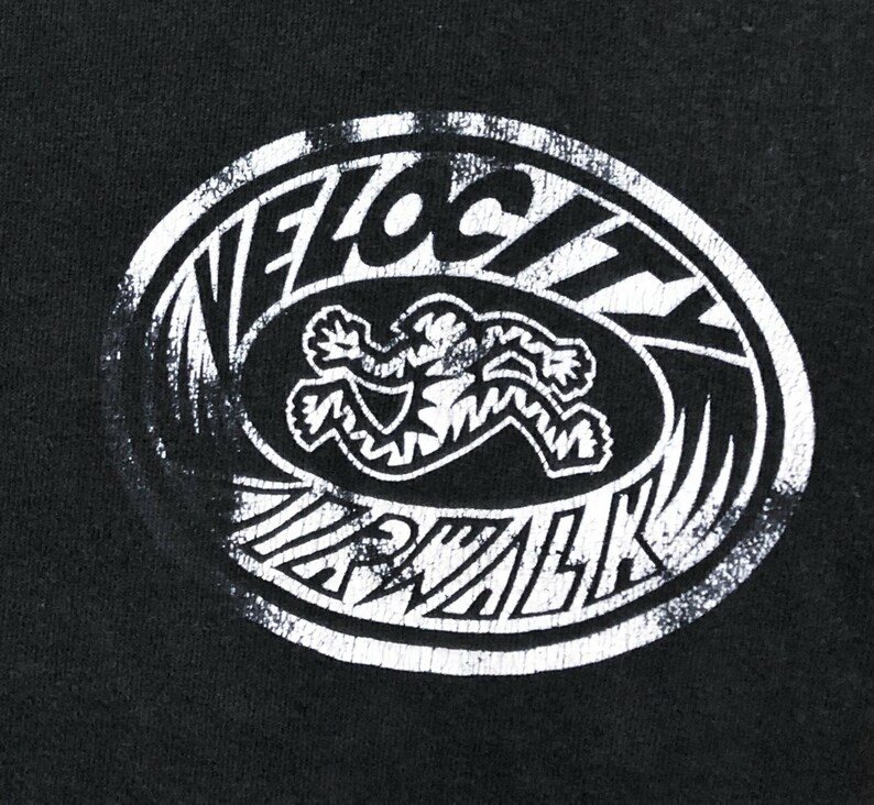 Op de afbeelding: Zwart t-shirt met een verweerde witte ovale afbeelding. De afbeelding toont de woorden "VELOCITY WALK" rond een gestileerde figuur in beweging. Het ontwerp heeft een vintage, versleten uitstraling.