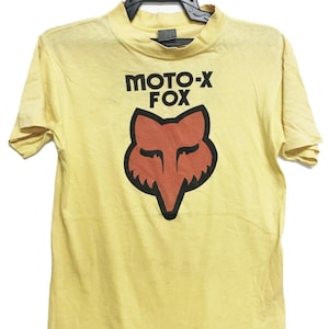 Peut inclure: T-shirt jaune à manches courtes avec le texte "MOTO-X FOX" en noir au-dessus d'un motif de tête de renard stylisée. La tête de renard est soulignée en noir avec un intérieur rouge-orange. Le t-shirt est suspendu à un cintre noir.