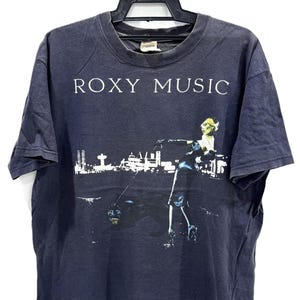Könnte beinhalten: Dunkelblaues T-Shirt mit dem weißen Schriftzug "ROXY MUSIC". Die Grafik zeigt eine Figur vor einer Stadtkulisse. Das T-Shirt hängt an einem schwarzen Kleiderbügel.