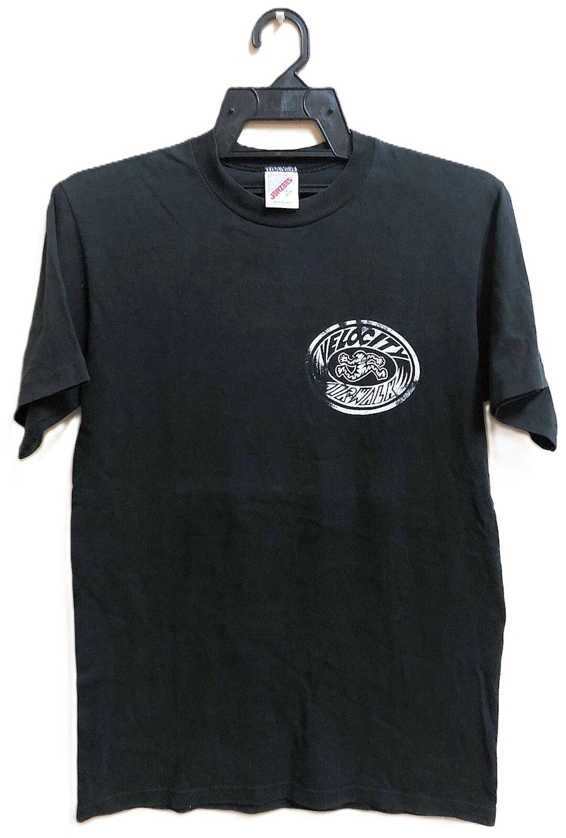 Op de afbeelding: Zwart T-shirt met een witte afbeelding. De afbeelding is een cirkelvormig ontwerp met het woord "VELOCITY" en een gestileerde surfer. Het shirt hangt aan een zwarte hanger.