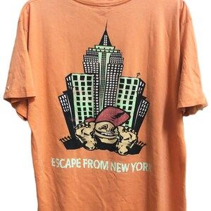 Peut inclure: Un t-shirt à manches courtes couleur pêche avec une impression graphique représentant une ligne d'horizon urbaine et un personnage de dessin animé au premier plan. Le texte "ESCAPE FROM NEW YORK" est imprimé en vert clair sous le motif.