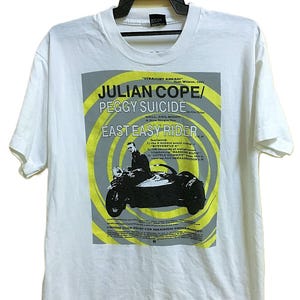 Può includere: T-shirt bianca con grafica dell'album "East Easy Rider" di Julian Cope e Peggy Suicide. Il design include un'immagine in bianco e nero di una moto e sidecar, con dettagli gialli e grigi. Il testo include il titolo dell'album e i titoli delle canzoni.