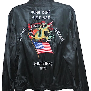 Puede incluir: Chaqueta de satén negro con bordado de dragón, bandera y texto. La parte trasera de la chaqueta presenta las palabras "HONG KONG", "VIET NAM", "JAPAN", "HAWAII", "PHILIPPINES" y el año "1971". La chaqueta tiene cierre de cremallera y está colgada de una percha negra.