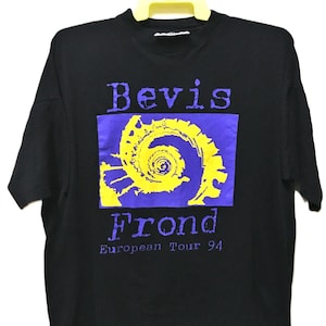 Può includere: T-shirt nero con la scritta "Bevis Frond" in viola, con un disegno a spirale giallo su sfondo viola. Il testo "European Tour 94" è in basso. La maglietta è appesa a una gruccia gialla.