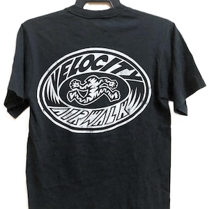 Op de afbeelding: Zwart t-shirt met een witte ovale afbeelding op de rug. De afbeelding toont de woorden "VELOCITY AIRWALK" rond een rennende figuur. Het shirt hangt aan een zwarte hanger.