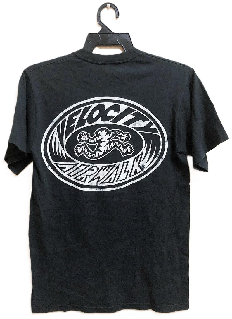 Op de afbeelding: Zwart t-shirt met een witte ovale afbeelding op de rug. De afbeelding toont de woorden "VELOCITY AIRWALK" rond een rennende figuur. Het shirt hangt aan een zwarte hanger.