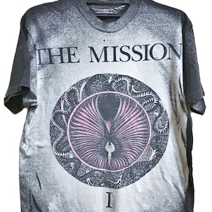 May include: A grey t-shirt featuring the text "THE MISSION" above a circular design with a pink and black graphic. The design includes a central figure and a surrounding pattern. The number "I" is below the design.