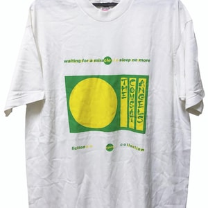 May include: White t-shirt featuring a green and yellow graphic. The design includes the text "waiting for a miracle sleep no more", "THE COMSAT ANGELS", "fiction", "pm", and "collection". The shirt is displayed on a black hanger.