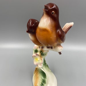 May include: A ceramic figurine of two brown birds perched on a branch with a white flower. The birds are painted with brown, orange, and white accents. The branch is painted green and has a white base.