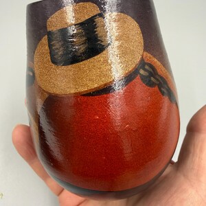 May include: A hand-painted, wooden tumbler with a design featuring a hat and a figure in shades of brown, gold, and red. The tumbler has a glossy finish and a rounded shape, suitable for beverages.