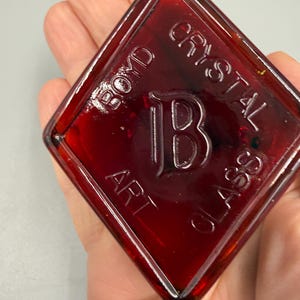 May include: A diamond-shaped, ruby-red glass piece with the embossed text "BOYD CRYSTAL ART GLASS" and a large letter "B" in the center. The glass has a glossy finish and is held in a hand.