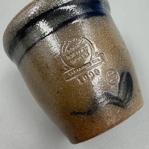 May include: A brown stoneware crock with a dark blue band around the rim. The crock has embossed text that reads "Rowe Pottery Works" and "1990". A black painted design is visible on the side.