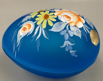 May include: A blue, egg-shaped decorative container with hand-painted floral designs in yellow, orange, and white. The flowers are accented with white leaves and a gold leaf detail. The container is a decorative item.