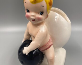 May include: A ceramic figurine of a cherub holding a black bowling ball. The cherub has blonde hair, blue eyes, and is wearing a pink diaper. The figurine is a vintage style and would make a great addition to any bowling enthusiast's collection.
