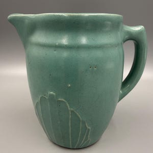 May include: A teal-colored ceramic pitcher with a handle and spout. The pitcher features a seashell design on the lower portion. The pitcher is a solid color with a matte finish.