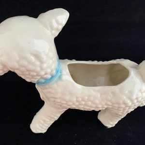 May include: White ceramic planter shaped like a lamb with a blue ribbon around its neck. The planter has a textured surface that resembles wool.