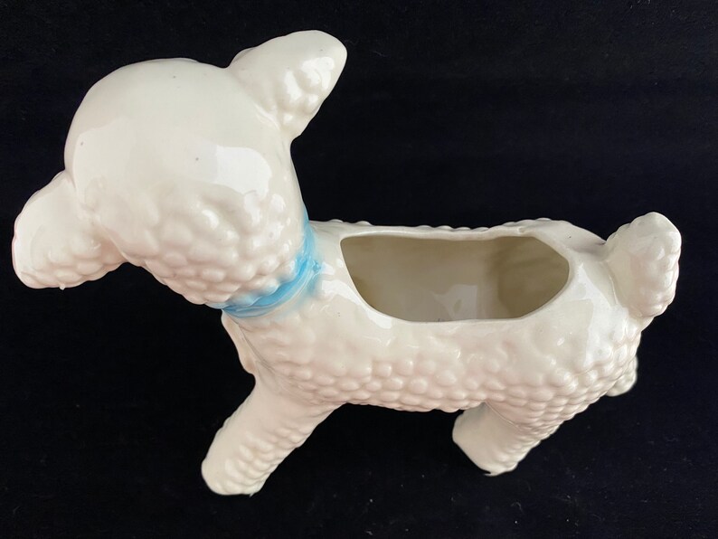 May include: White ceramic planter shaped like a lamb with a blue ribbon around its neck. The planter has a textured surface that resembles wool.
