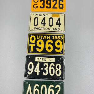 May include: Five vintage license plates from different states. The top plate is orange with black text and reads "TEXAS 53 G 3926". The second plate is cream with black text and reads "MAINE 0404 VACATIONLAND". The third plate is black with yellow text and reads "UTAH 1953 Q 969". The fourth plate is black with white text and reads "MASS 53 94-368". The bottom plate is green with white text and reads "A6062 VERMONT 53".