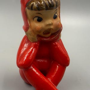 May include: A glossy ceramic figurine of a seated figure wearing a bright red one-piece suit and a matching red cap with a pom-pom. The figure has brown hair, rosy cheeks, and an open-mouthed expression, with hands resting near the chin.