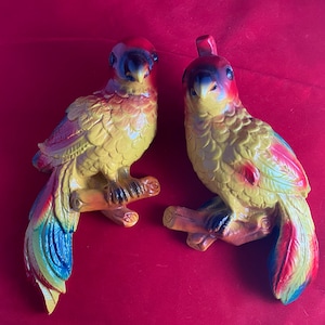 May include: Two ceramic parrot figurines with yellow, red, and blue paint. The parrots are perched on branches and have detailed feathers.