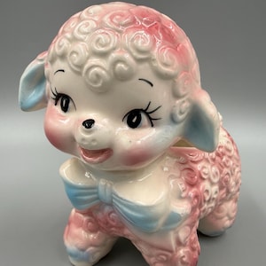 May include: A ceramic lamb planter featuring a glossy white finish with hand-painted pink curls and light blue accents on the ears and bow. The lamb has large black eyes with long eyelashes, a small black nose, and a smiling mouth with pink cheeks.