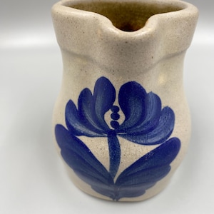 May include: A small, cream-colored ceramic pitcher with a blue floral design. The pitcher has a rounded body and a small spout.