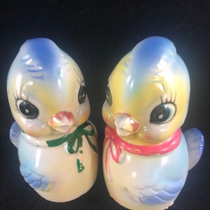 May include: A pair of ceramic salt and pepper shakers shaped like birds. The birds are yellow with blue wings and have large, black eyes. One bird has a green bow and the other has a pink bow.