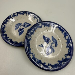 May include: Two small, round ceramic plates. The plates have a speckled, off-white base with a blue floral design in the center. A blue, decorative border encircles the rim. The plates are likely for serving small portions or as decorative pieces.