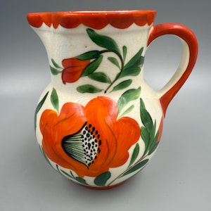 May include: A white ceramic pitcher with a red rim and handle. The pitcher is decorated with hand-painted orange flowers and green leaves.