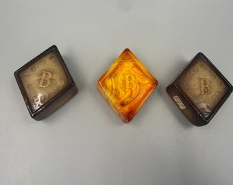 May include: Three diamond-shaped glass paperweights with a swirled finish. Two are brown and one is orange. Each features a central letter B and the embossed text Boyd Crystal Art Glass. The items are arranged in a horizontal row on a flat surface.