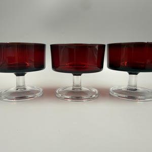 May include: Three matching dessert bowls made of deep red glass. Each bowl features a wide, cylindrical basin supported by a short, clear glass stem and a circular, flat clear glass base, providing a stable foundation for the colored glass top.