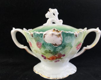 May include: A white porcelain sugar bowl with a lid, featuring a green and white floral design with gold accents. The bowl has two handles and a scalloped edge.