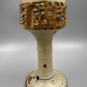 May include: A ceramic chalice with a speckled cream glaze and a tall, slender stem. The upper bowl features a cut-out grid pattern with brown glaze accents, small blue painted cross motifs, and tiny white raised dots across the textured surface.