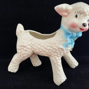 May include: White ceramic planter shaped like a lamb with a blue bow. The lamb has a textured surface and a small opening on its back for planting.