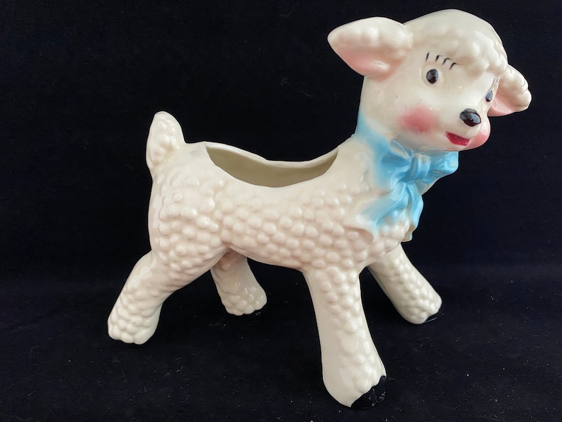 May include: White ceramic planter shaped like a lamb with a blue bow. The lamb has a textured surface and a small opening on its back for planting.