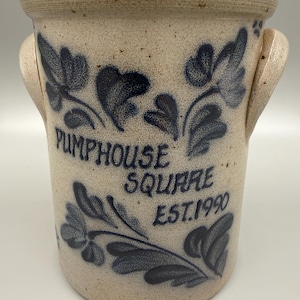 May include: A stoneware crock with a light beige base and dark blue floral designs. The crock has two handles and the words "PUMPHOUSE SQUARE EST.1990" are printed on the front. This vintage piece is ideal for kitchen decor.