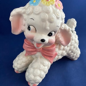 May include: A white ceramic poodle figurine with pink ears, a pink bow, and a flower crown with blue, yellow, and pink flowers.