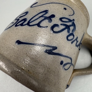 May include: A stoneware mug with a handle, featuring the words "Salt Fork" in dark blue script. The mug has a speckled, light brown finish. The mug is angled, showing the side and handle.
