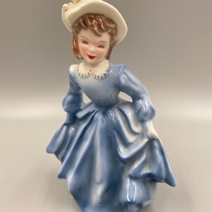 May include: A porcelain figurine of a girl wearing a blue dress and a white hat with a brown ribbon. She has brown hair and a painted smile.