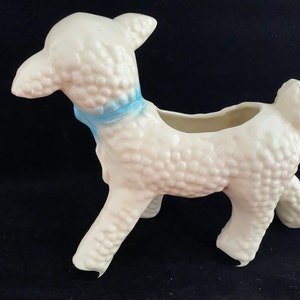 May include: White ceramic lamb planter with a blue ribbon around its neck. The lamb has a textured surface and is standing on all four legs. The planter has a small opening on the top for planting.