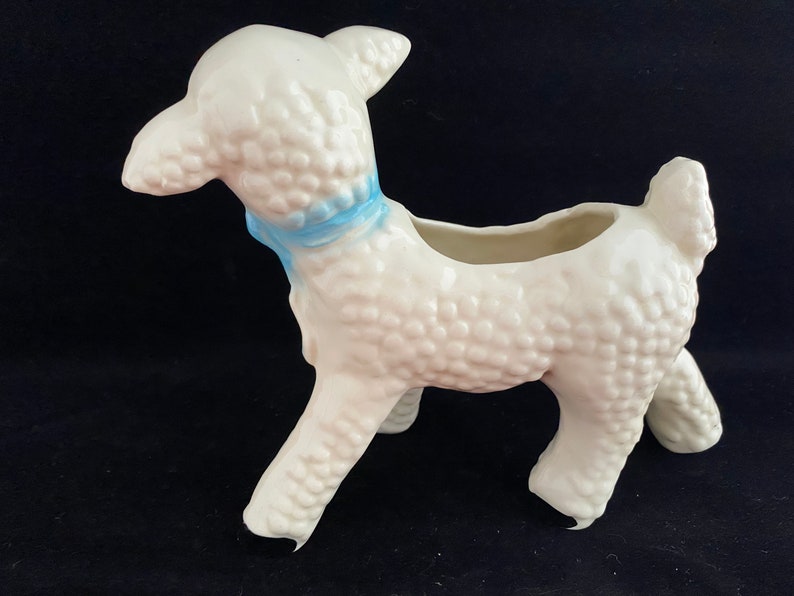 May include: White ceramic lamb planter with a blue ribbon around its neck. The lamb has a textured surface and is standing on all four legs. The planter has a small opening on the top for planting.