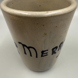 May include: A beige stoneware crock with a wide opening and a tapered body. The word "MERRY" is painted in dark blue on the side. The crock has a rustic, aged appearance, suggesting it is vintage or antique.